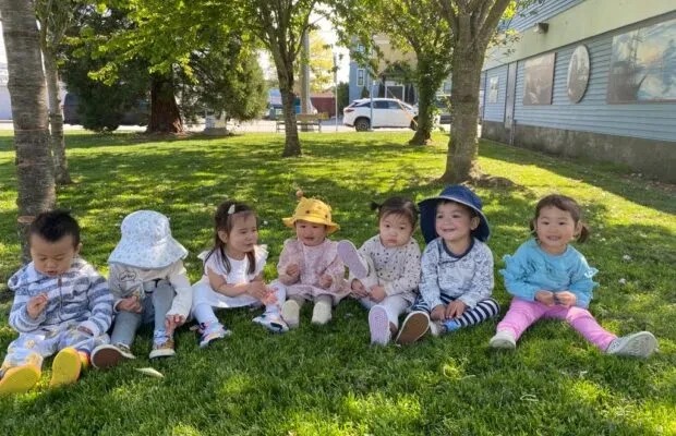 Seven toddlers sitting in a row on the grass under the trees, some in sun hats