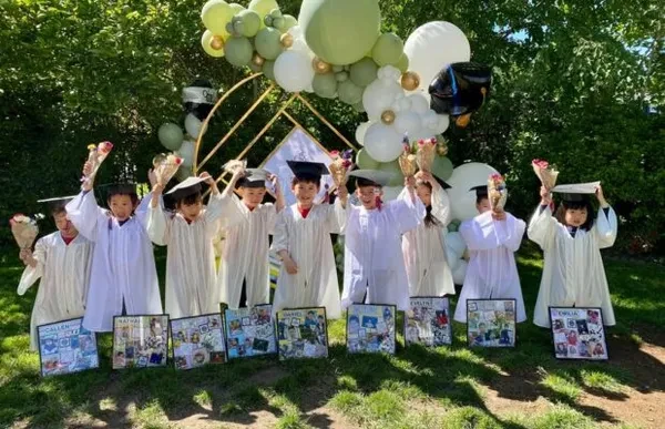 Children in white graduation gowns and caps holding up flowers in front of a balloon arch