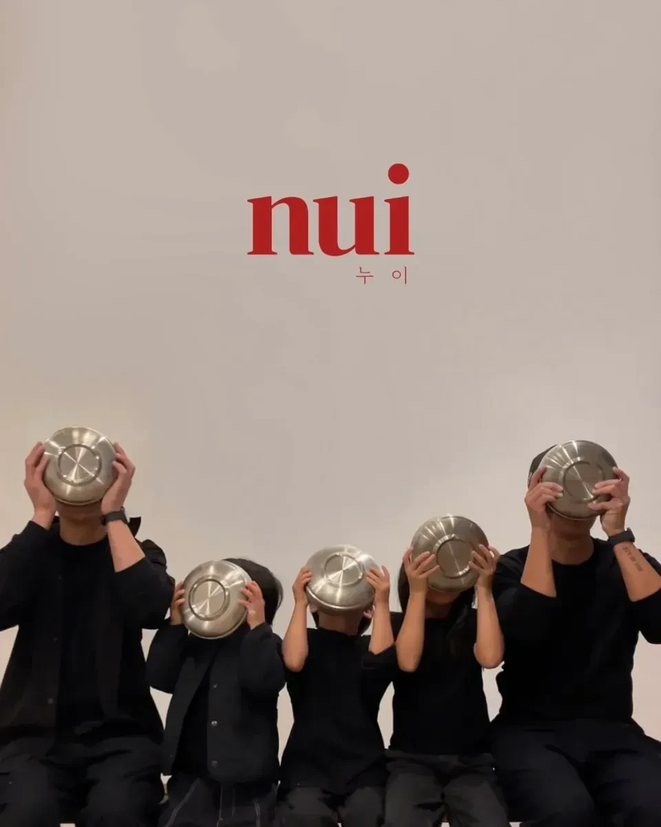 Five people in black sitting in a row, each holding a steel bowl over their face, under a red Nui logo on a white wall