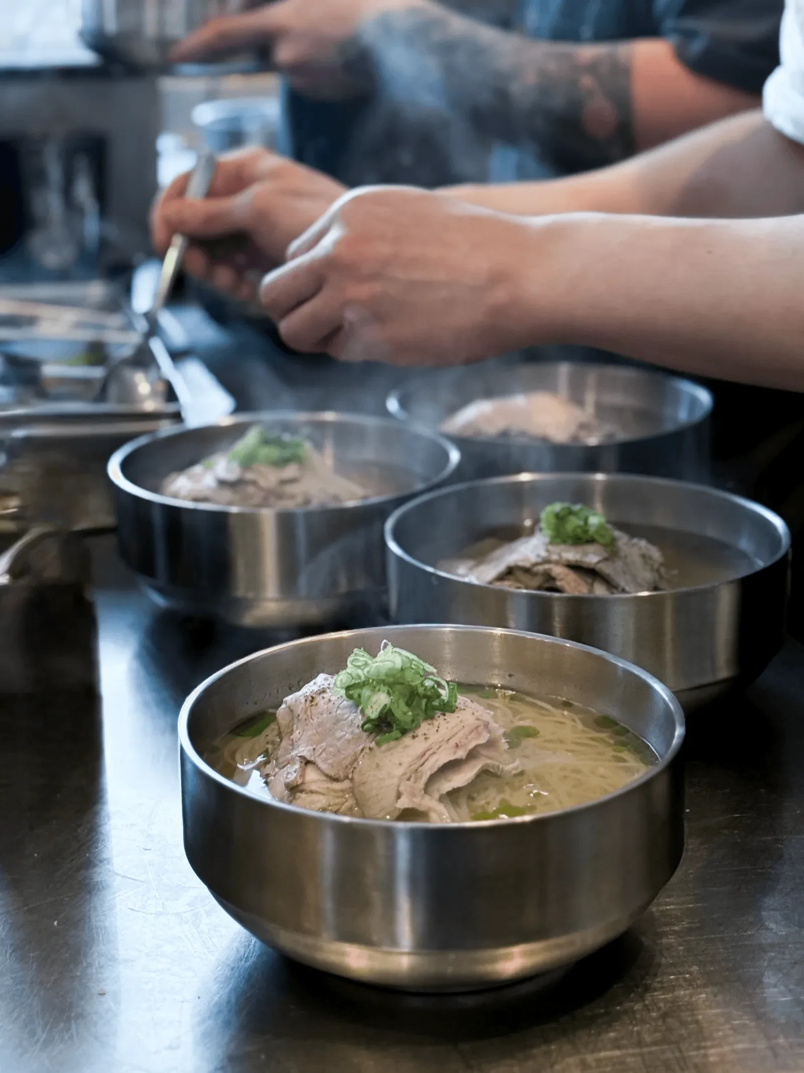 Steaming steel bowls of noodle soup topped with sliced meat and green onion, lined up on a counter as cooks work behind them