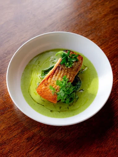 A seared salmon fillet topped with fresh herbs in a bright green sauce, served in a white bowl on a wooden table