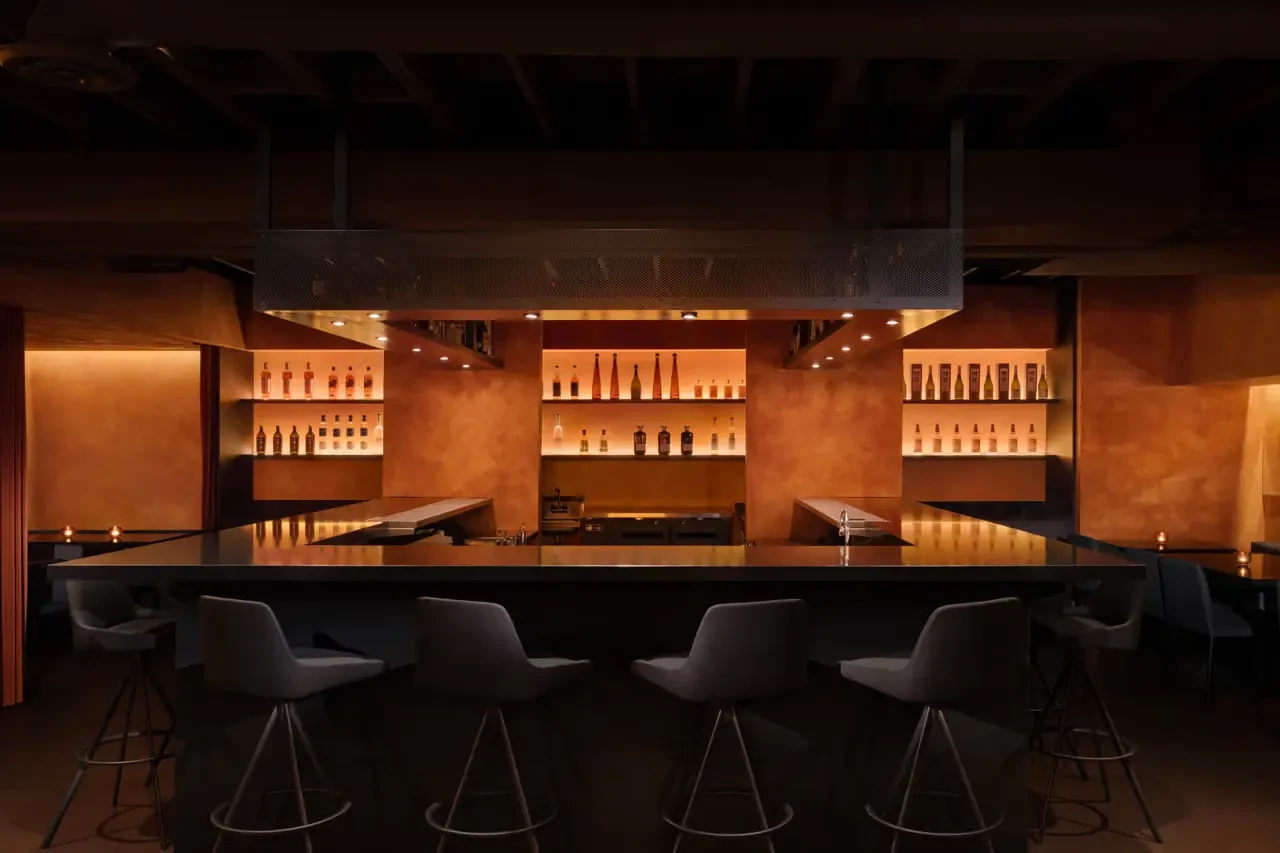 The dimly lit AMA Raw Bar: grey stools around a dark bar, with glowing shelves of bottles set into warm orange walls