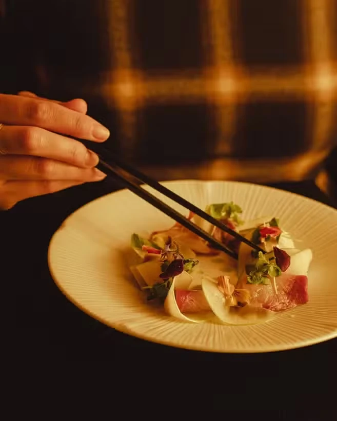 Chopsticks lifting from a plate of thinly sliced raw fish with shaved radish, herbs and edible flowers