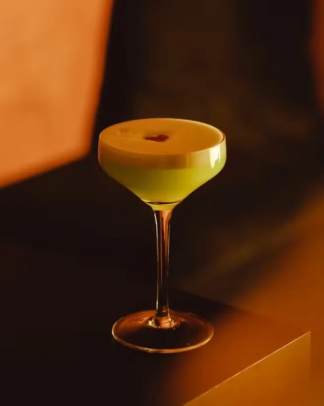 A frothy pale green cocktail in a coupe glass on a ledge in warm amber light