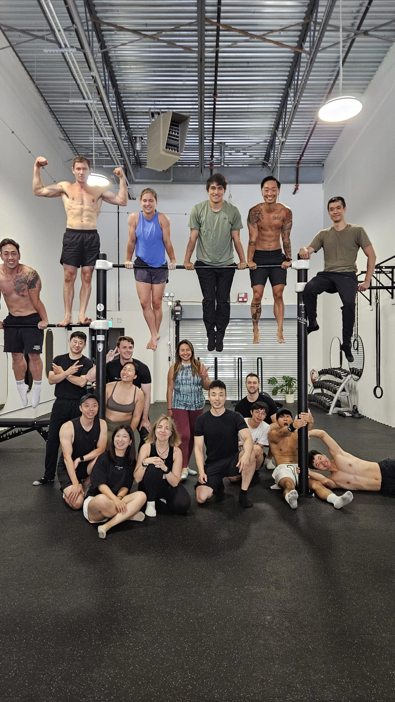 A big group of Calibar members posing around a pull-up bar, some hanging from it, some standing on it and others sitting on the mat