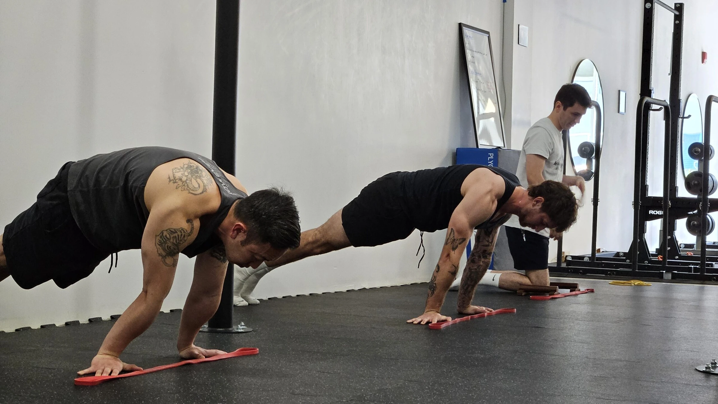 Two members in a deep plank with their hands on red resistance bands while a coach watches from behind