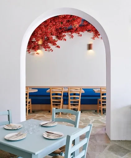 A white archway hung with red flowers, looking past a pale blue table and chairs into a dining room with wooden chairs and a blue banquette
