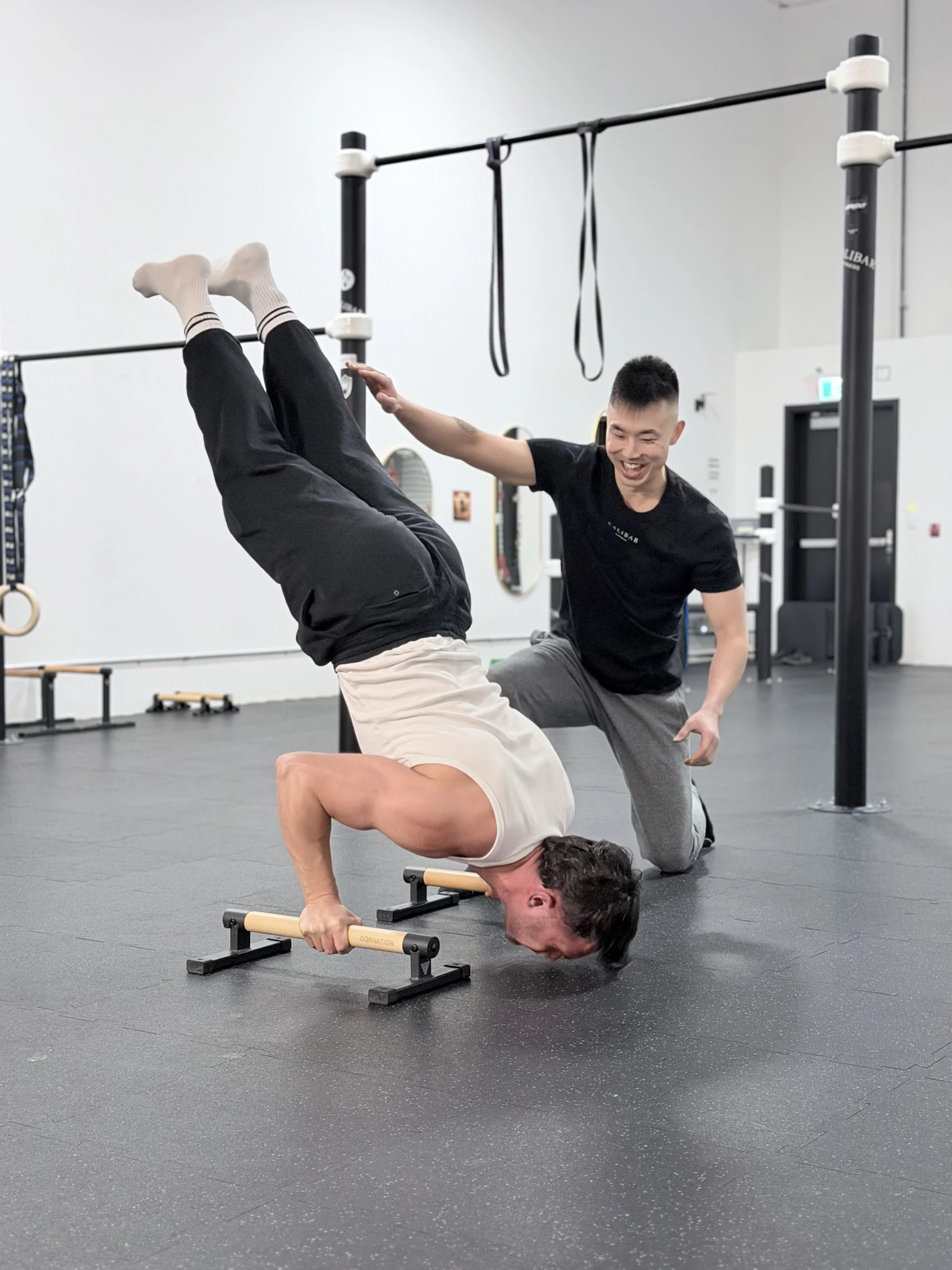 A coach in a black Calibar shirt kneels and laughs as a member in a white tank top holds a headstand on wooden parallettes